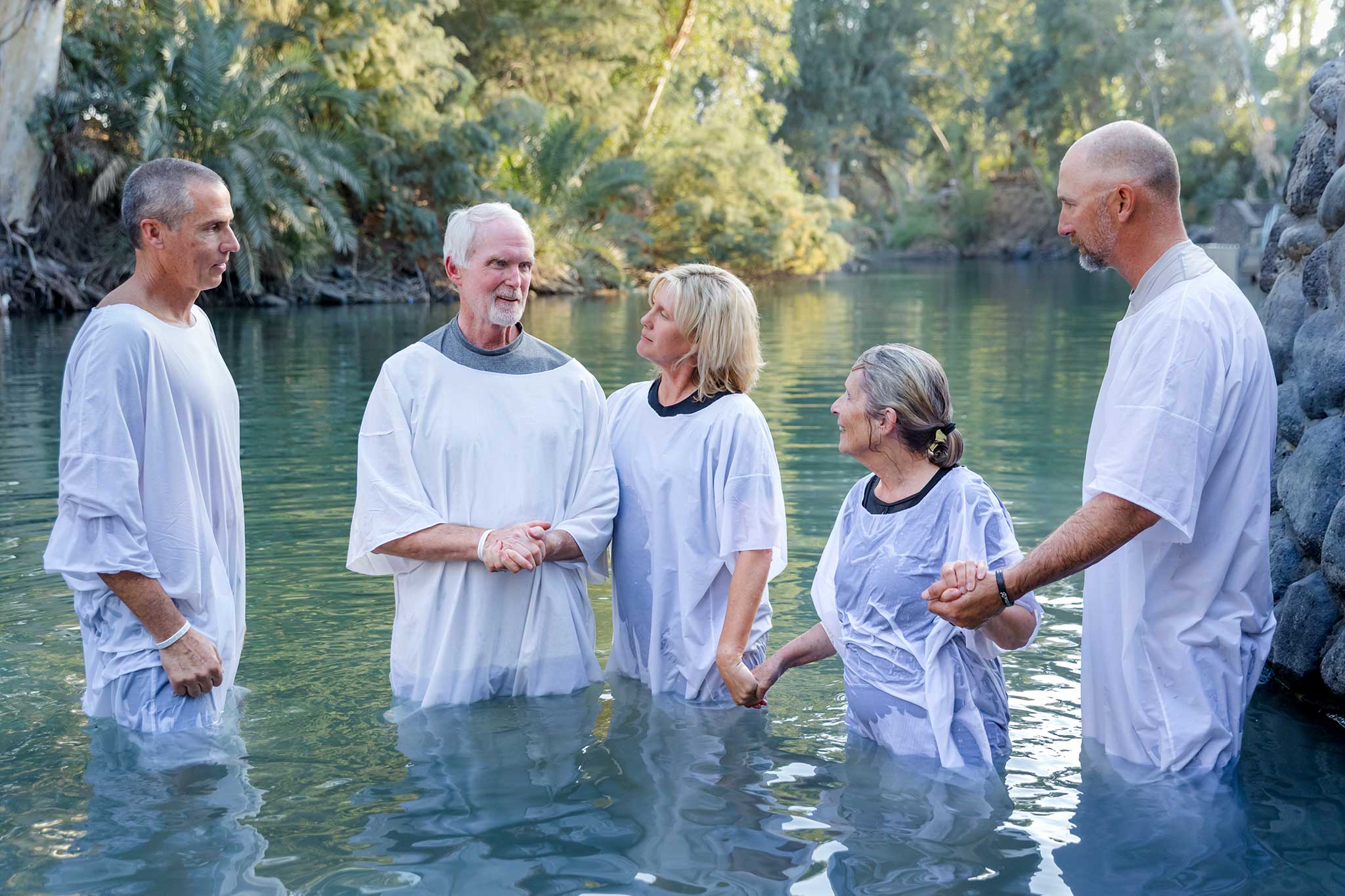 Five people in white baptism robes stand together in the Jordan River surrounded by lush greenery.