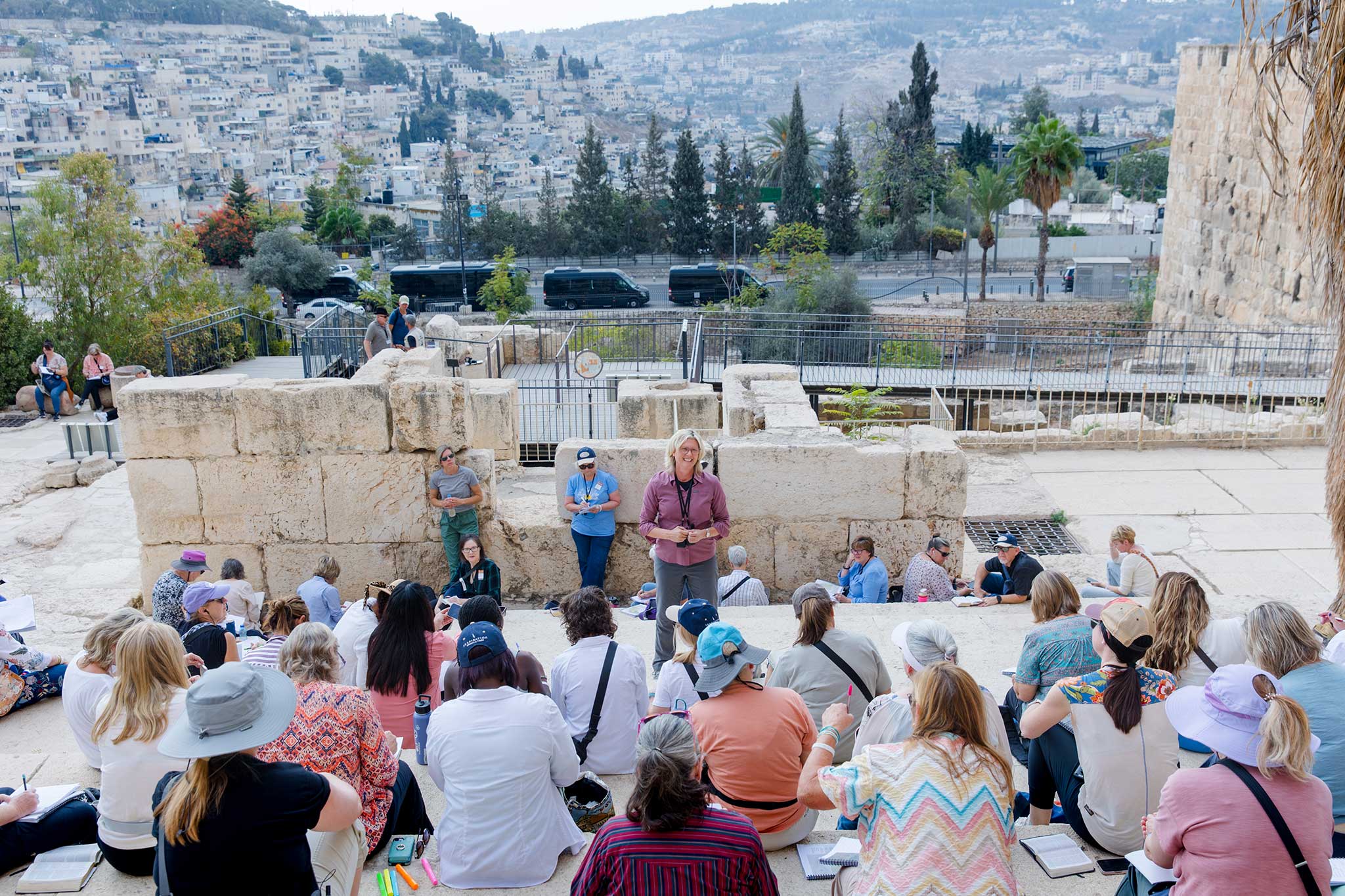 A group gathers around a speaker among ancient stone ruins, with Jerusalem visible in the background.