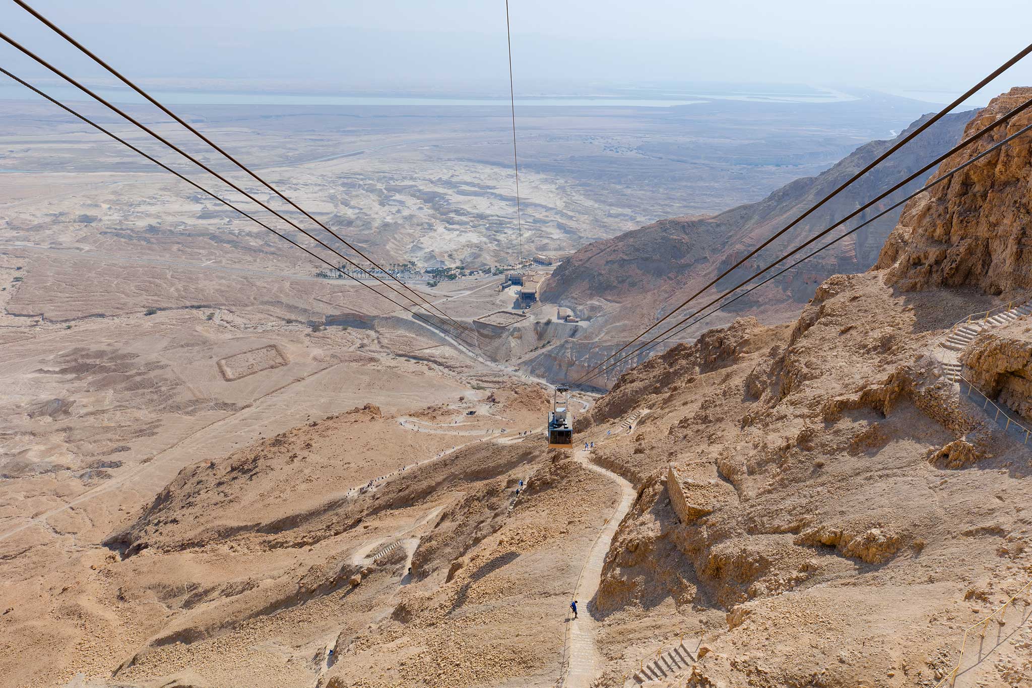 A cable car descends from Masada over the rugged Judean Desert with the Dead Sea visible in the distance.