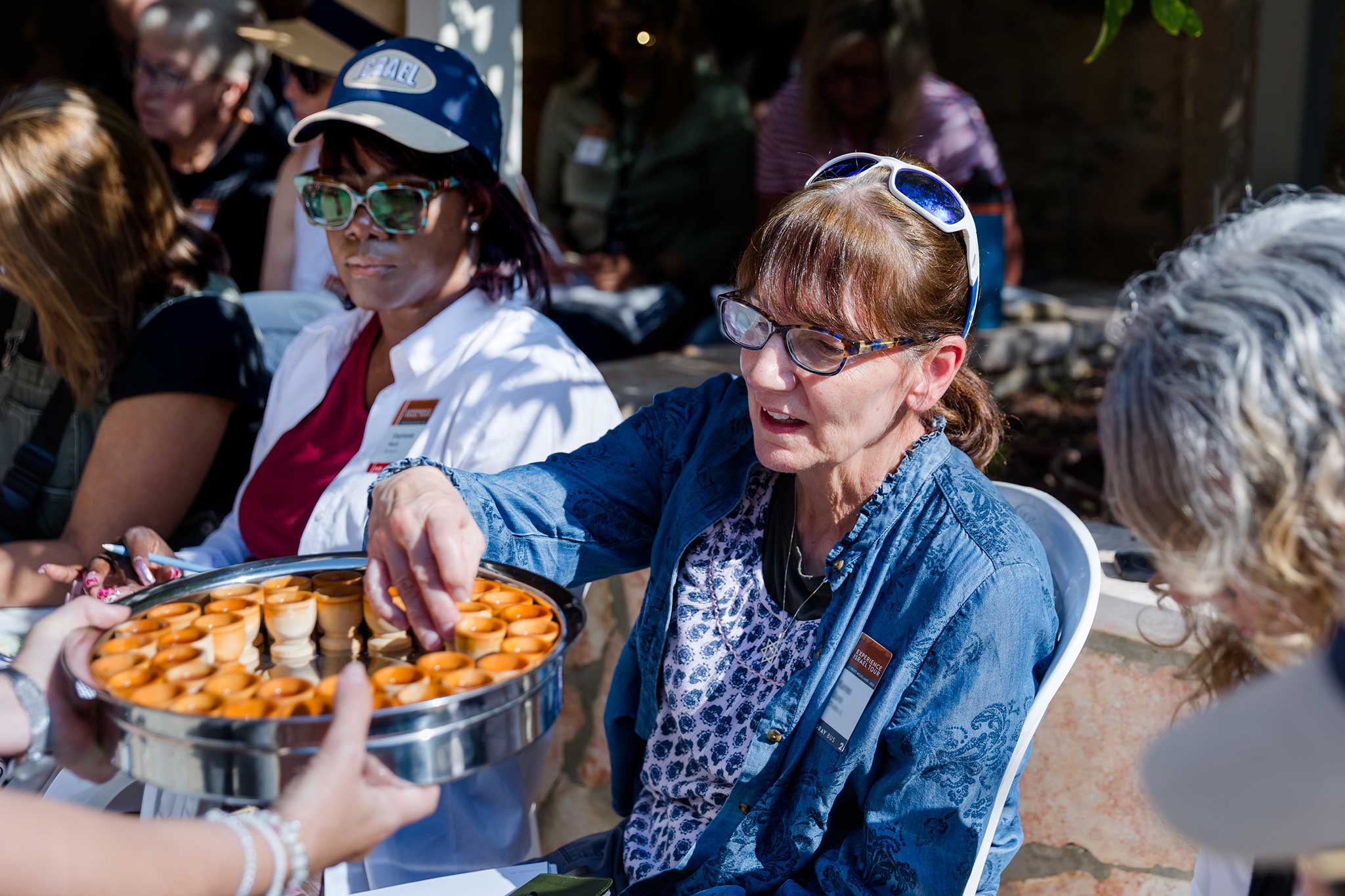 A traveler reaches for a communion cup from a tray while seated with a group outdoors.