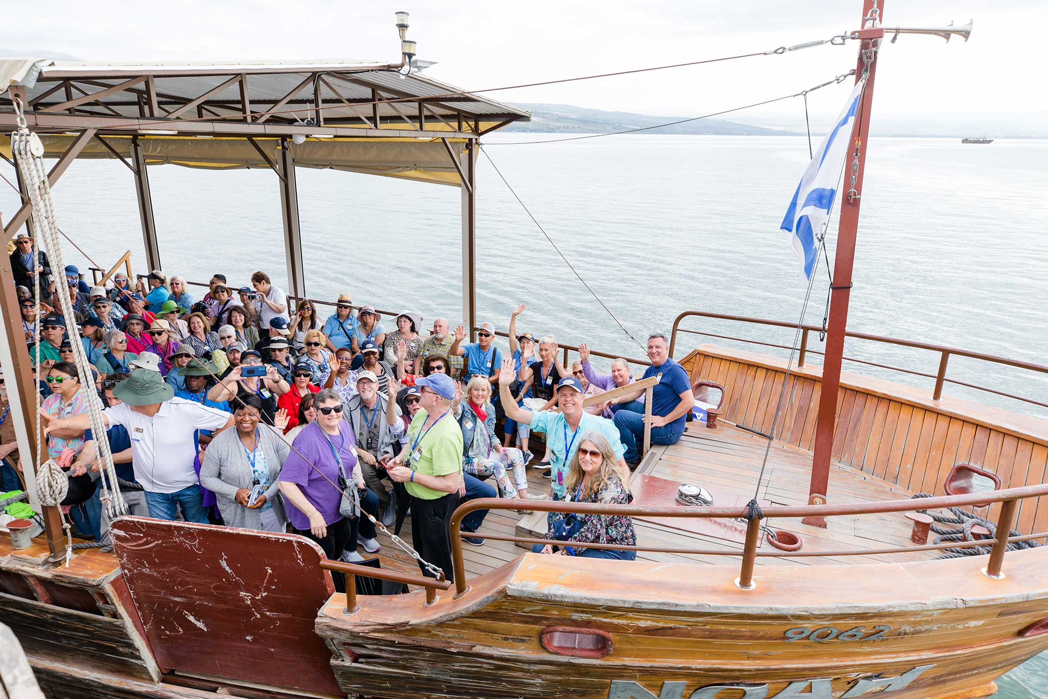 Travelers gather aboard a wooden boat on the Sea of Galilee, smiling and waving beneath an Israeli flag with peaceful water behind them.