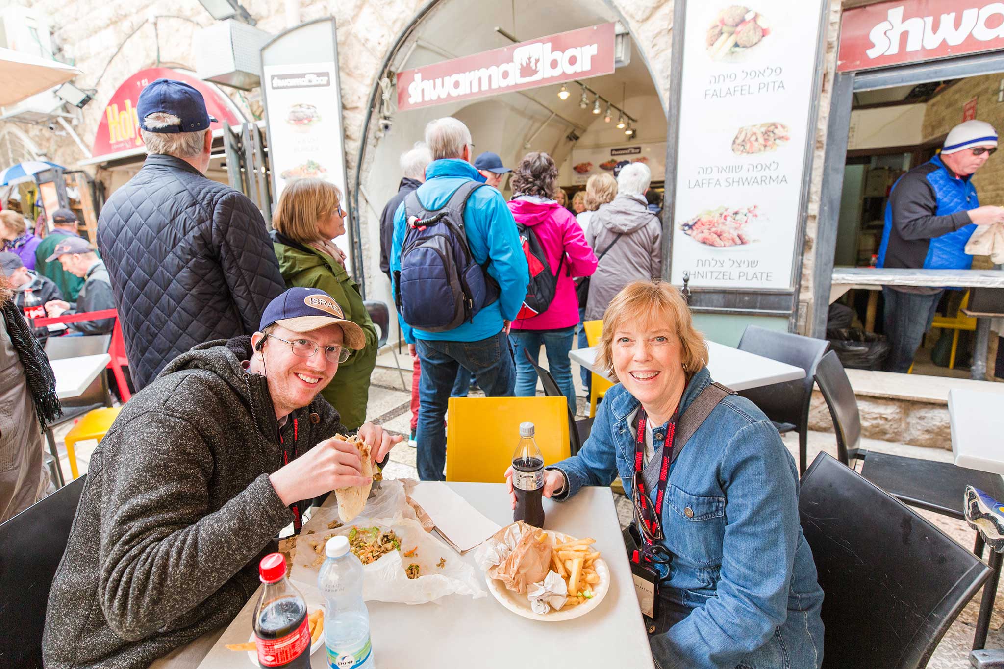 Christian travelers enjoy falafel, shawarma, and fries at an outdoor cafe, smiling together during a lively group stop in Israel.