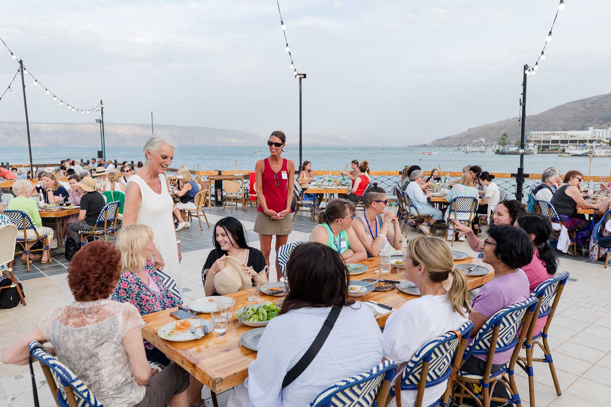 Women enjoy an outdoor waterfront meal together, sharing conversation at tables overlooking the Sea of Galilee and surrounding hills.