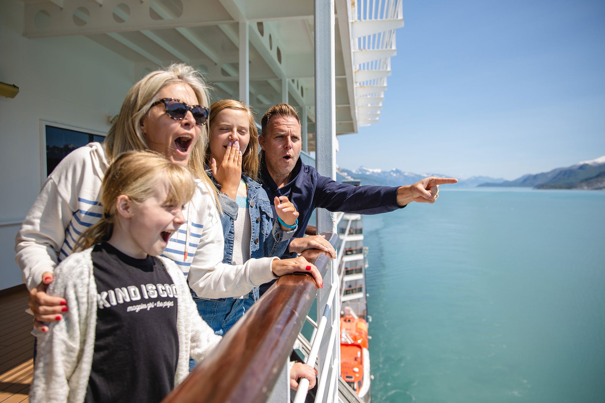 A family stands on the deck of a cruise ship, smiling and pointing toward Alaska's scenic coastline and mountains as they take in the breathtaking view.