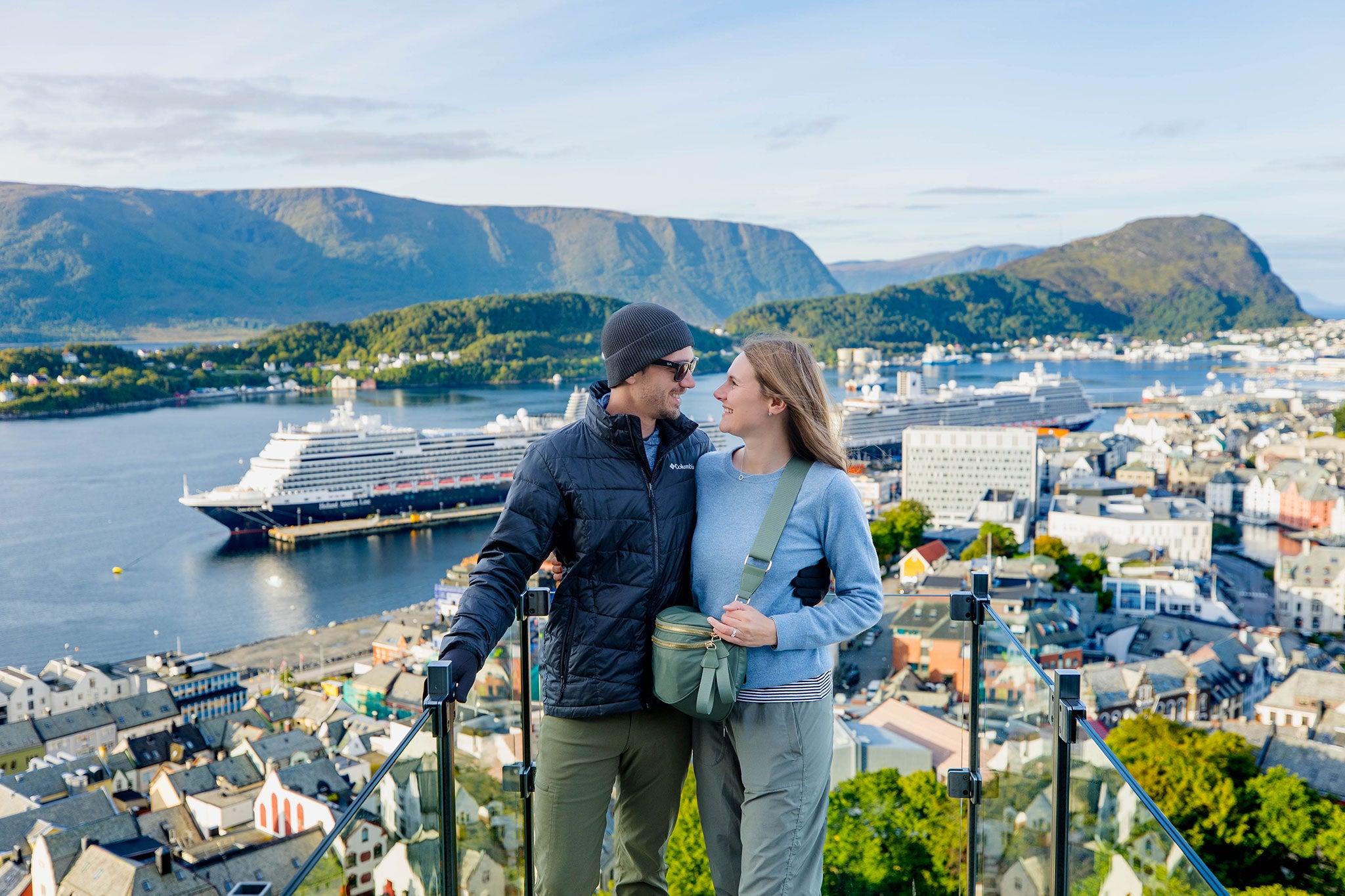 Smiling couple at viewpoint overlooking coastal town, fjord, mountains and cruise ship.