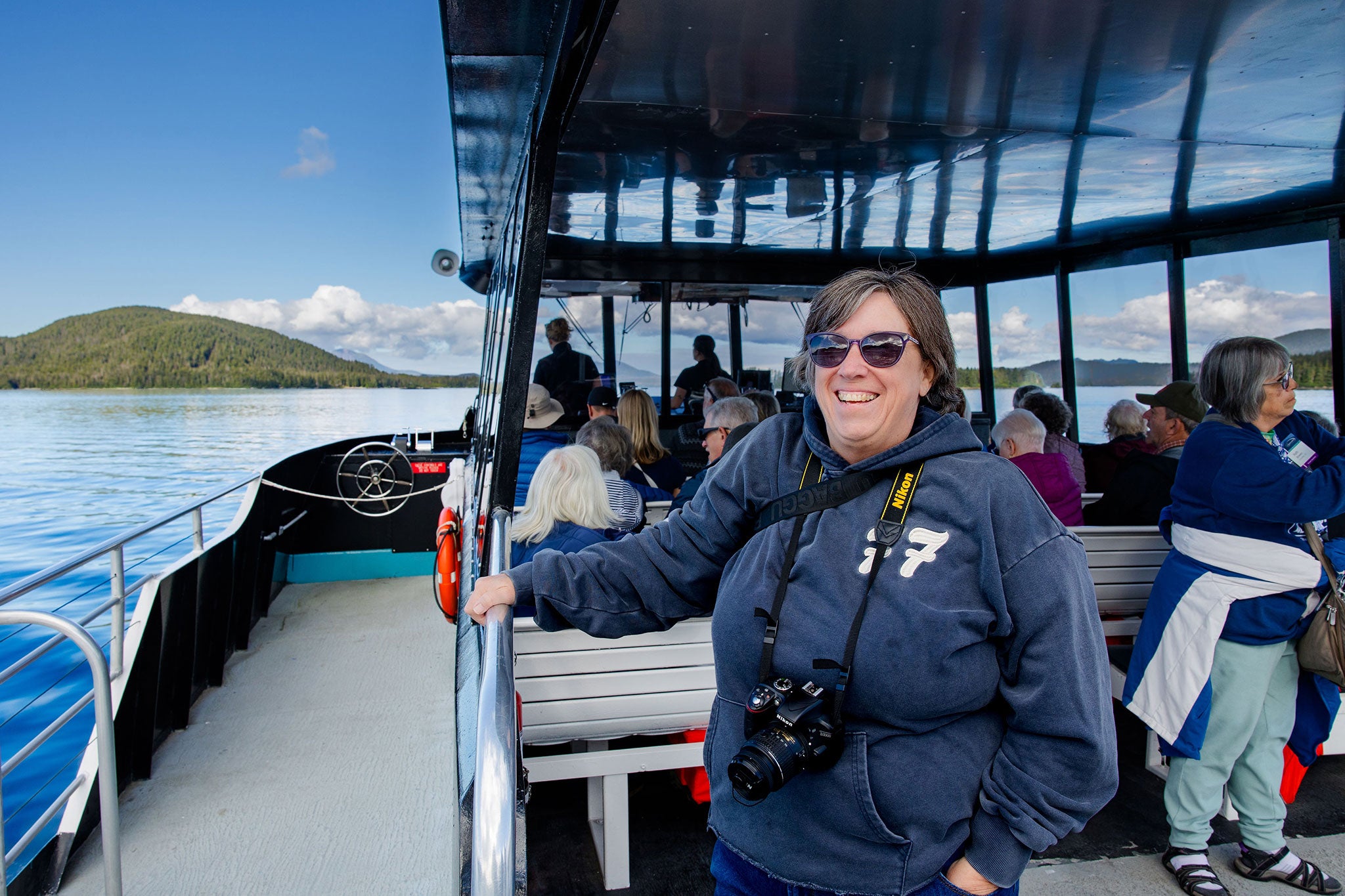 Woman with sunglasses and camera on boat deck, passengers seated behind
