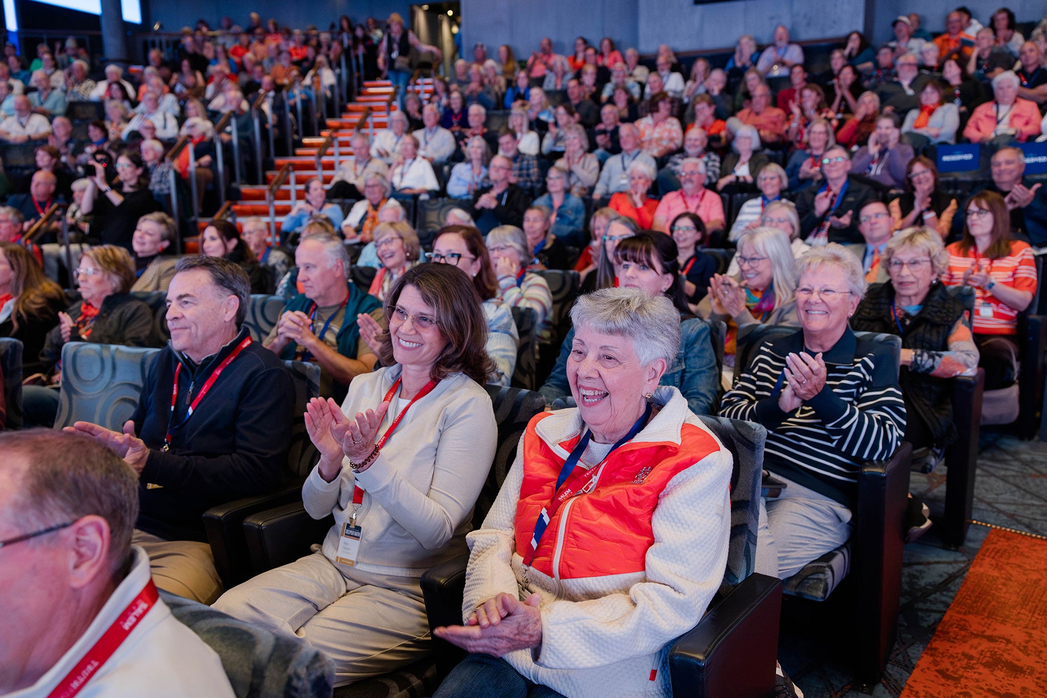 Photograph of a large seated audience in an auditorium, many applauding and wearing red lanyards.
