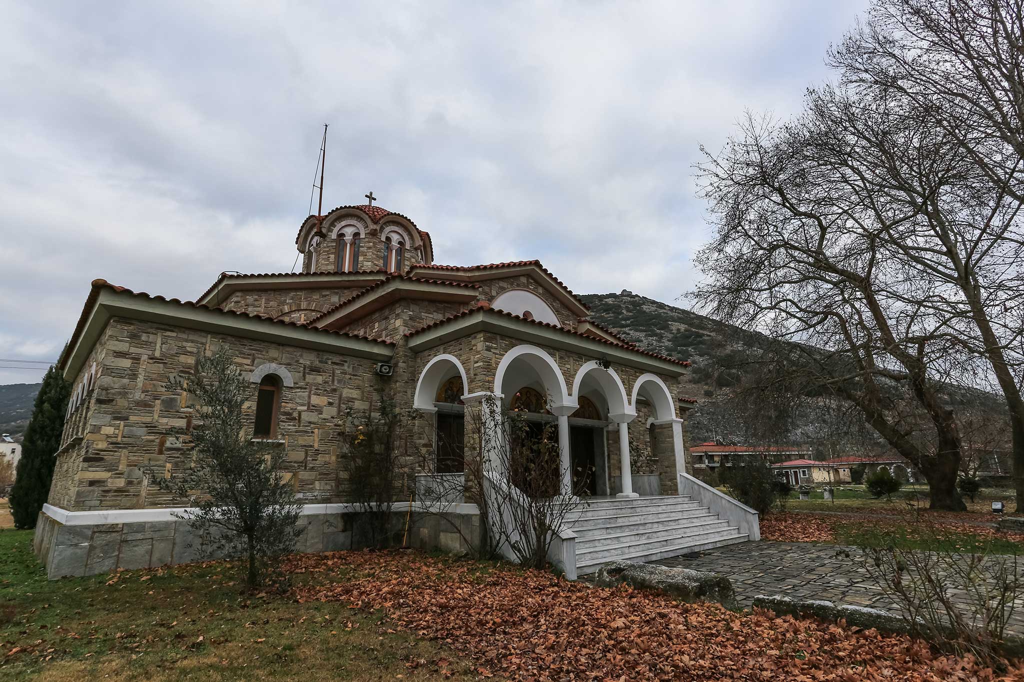 Stone church with arched porch, fallen leaves and bare trees under cloudy sky