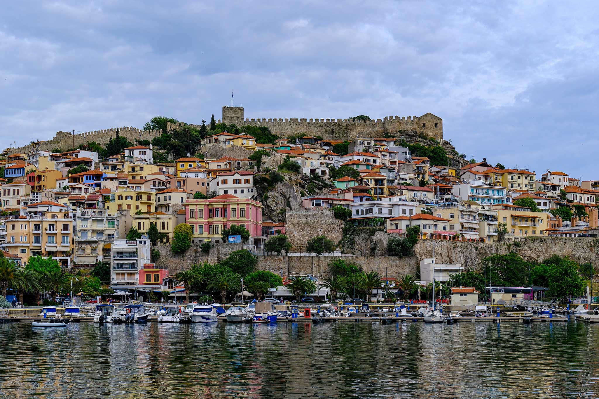 Colorful hillside town with a hilltop fortress reflected in calm water under cloudy sky