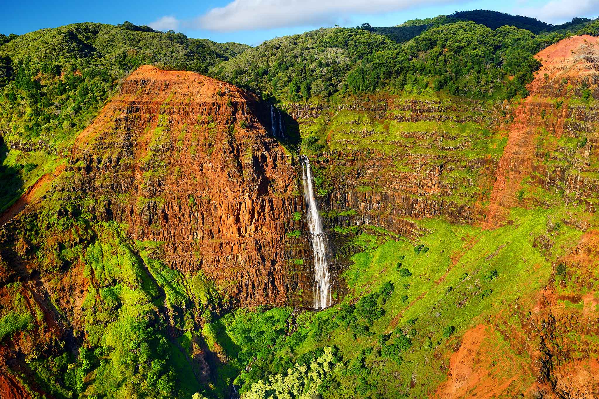Waterfall drops between red cliffs into a lush green canyon.