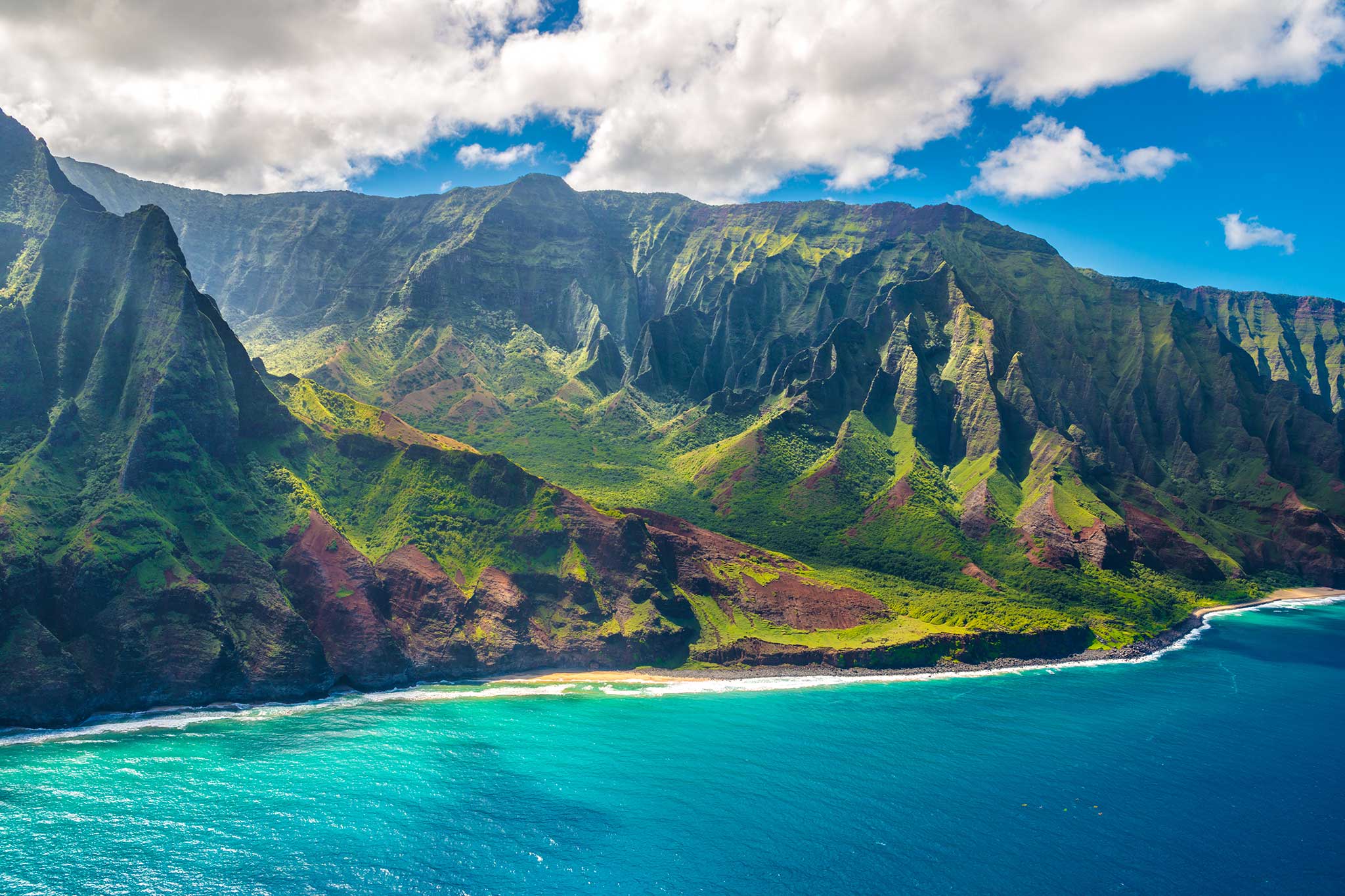 Turquoise ocean and sandy beach below lush green mountains under bright clouds