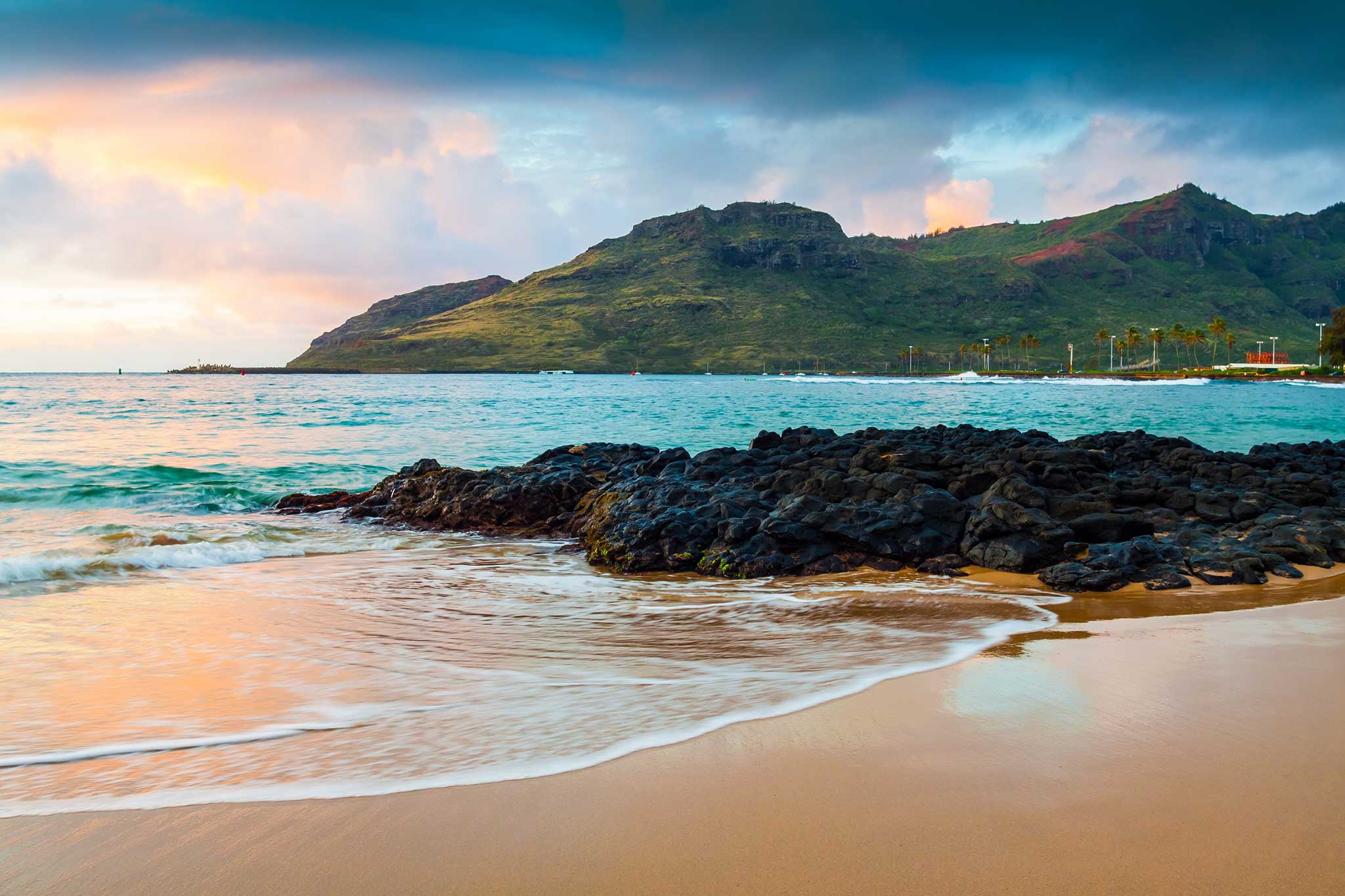 Sunset beach with gentle waves, dark rocks and green mountains in the distance.
