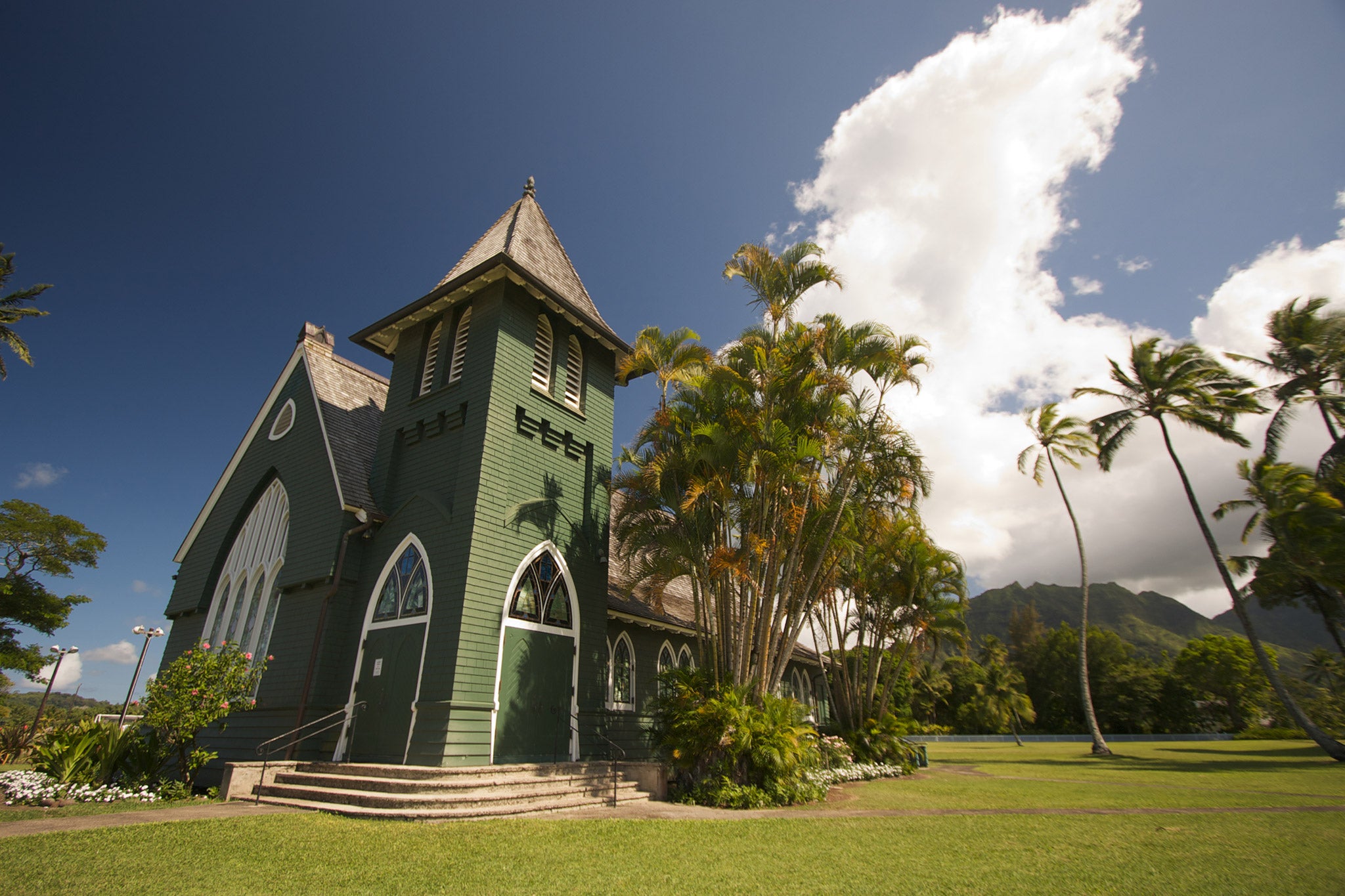 Photograph of a green church on a sunny lawn with palm trees and a large cloud