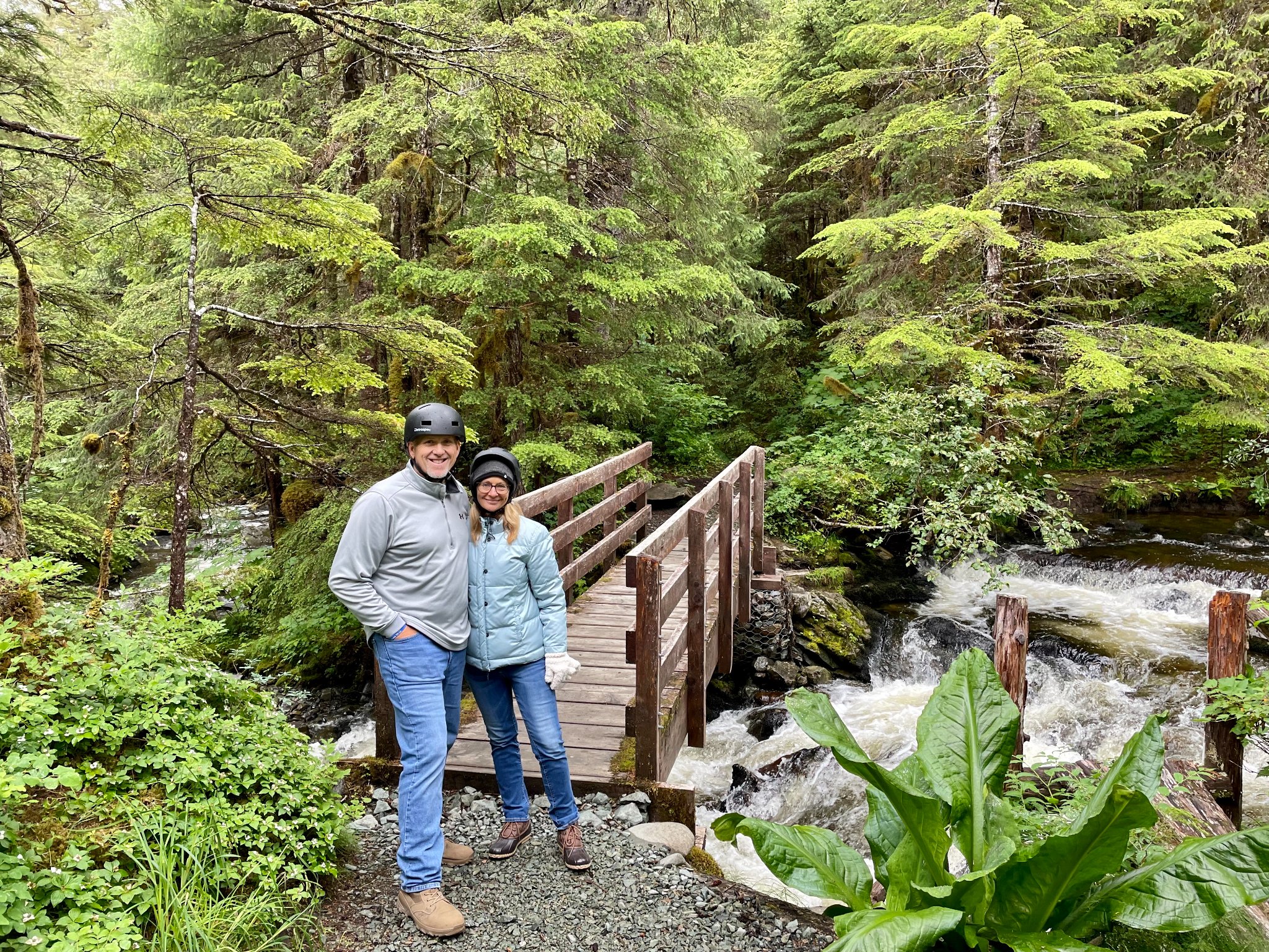A couple standing together with a small wooden bridge, river and tall trees in the background.