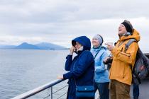 Two women and a man wearing windbreaker jackets and headbands, carrying various bags, cameras and coffee cups, stand at a cruise ship’s railing, laughing off-camera with the icy blue ocean and mountains in the background.