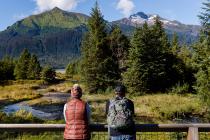 Two people dressed for cool weather enjoy a scenic mountain view from a wooden overlook in Alaska.