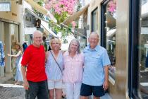 Four smiling travelers pose together on a sunny cobblestone street lined with shops and flowers.