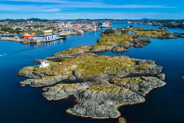 Aerial view of small rocky islands with a white lighthouse near Haugesund, Norway, surrounded by calm blue water and distant cityscape