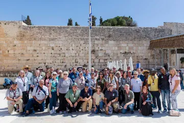 A group of travelers poses together in front of the Western Wall in Jerusalem beneath a bright blue sky and Israeli flag.
