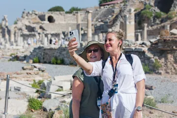 Two travelers smile for a selfie while exploring the ancient ruins of Ephesus, with weathered stone columns and historic structures in the background.