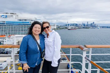Photo of two women smiling on ship deck, cruise liner and city skyline behind.