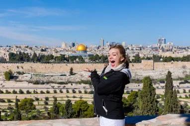 A smiling woman traveler stands at a scenic overlook in Jerusalem, gesturing toward the Old City skyline with the golden Dome of the Rock visible in the distance under a clear blue sky.