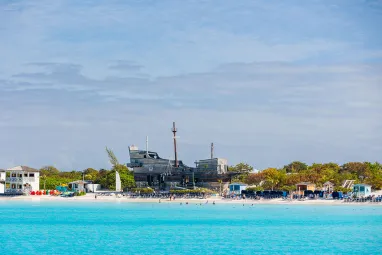 A bright turquoise shoreline with calm ocean water in the foreground, beach chairs and small colorful buildings along the sand, and a large wooden pirate ship structure set among palm trees under a blue, lightly clouded sky.