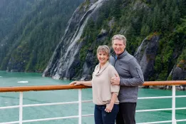 Smiling couple on a cruise deck with emerald fjord waters, rocky cliffs, and waterfalls in the background.