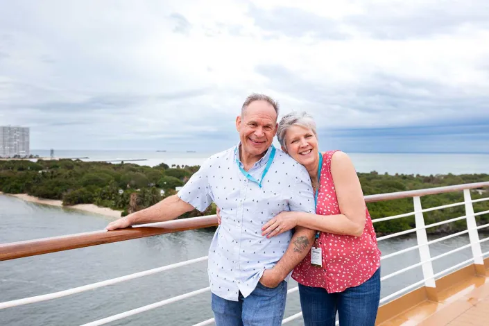 A smiling older couple embracing each other on a cruise ship deck, with a coastal landscape and the sea in the background under a cloudy sky.