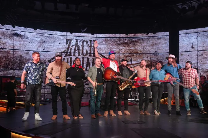 Zach Williams with a group of musicians onstage during opening night, taking a bow with guitars and other instruments in front of a rustic backdrop.