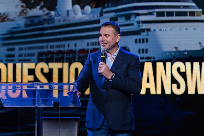 Amir Tsarfati stands onstage behind a clear podium, holding a microphone as he speaks during a Behold Israel Alaska cruise. Behind him, a large screen shows a cruise ship set against dramatic coastal mountains with the words “Questions & Answers.”