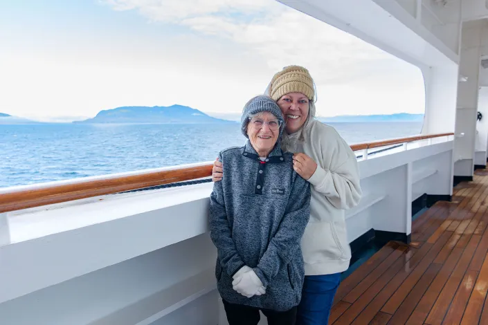 Smiling woman and elderly companion bundled in knit hats, gloves and warm layers on a cruise ship deck. They stand beside the railing with ocean views and a distant island in the background, enjoying a cool day at sea.