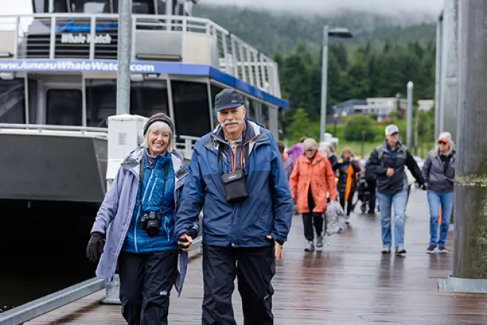 A group of travelers walks along a waterfront dock during an Alaska shore excursion. Two smiling travelers in outdoor jackets lead the way, with tour boats and forested hills in the background.