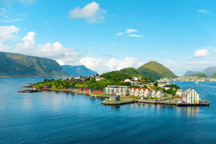 Alesund, a scenic coastal village along a Norwegian fjord, surrounded by green hills, mountains and calm blue water beneath a bright sky with scattered clouds. Colorful waterfront buildings line the shoreline, creating a picturesque view of Norway’s rugged beauty.