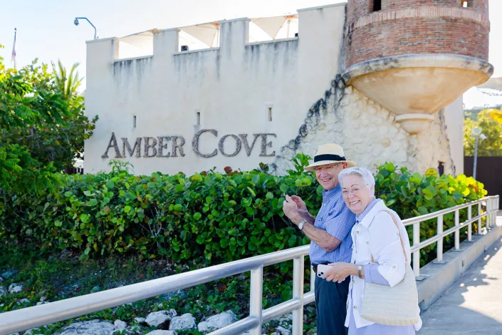 Two smiling travelers walk along a pathway in front of the Amber Cove entrance sign in the Dominican Republic. Surrounded by tropical greenery and historic-style architecture, they enjoy a sunny day exploring the cruise port.