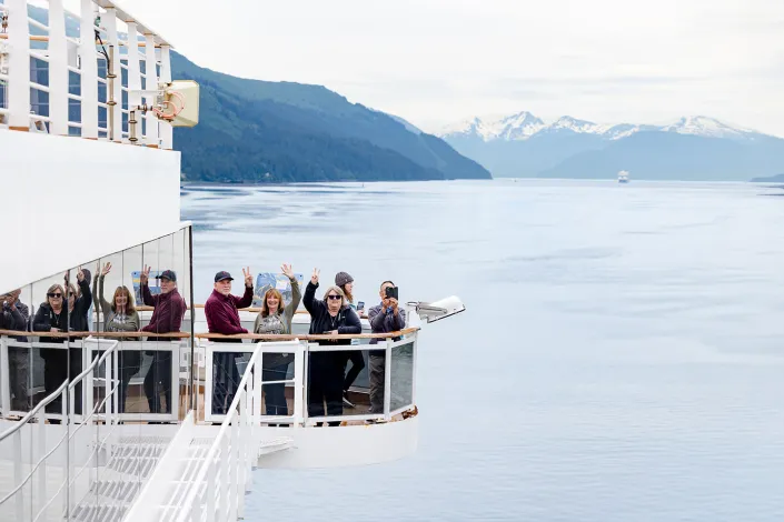 Travelers wave from the balcony of a cruise ship as it sails through calm Alaskan waters, surrounded by forested hills and snowcapped mountains.