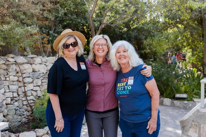 Kristi poses with two women, their arms around each other, in a leafy outdoor setting.