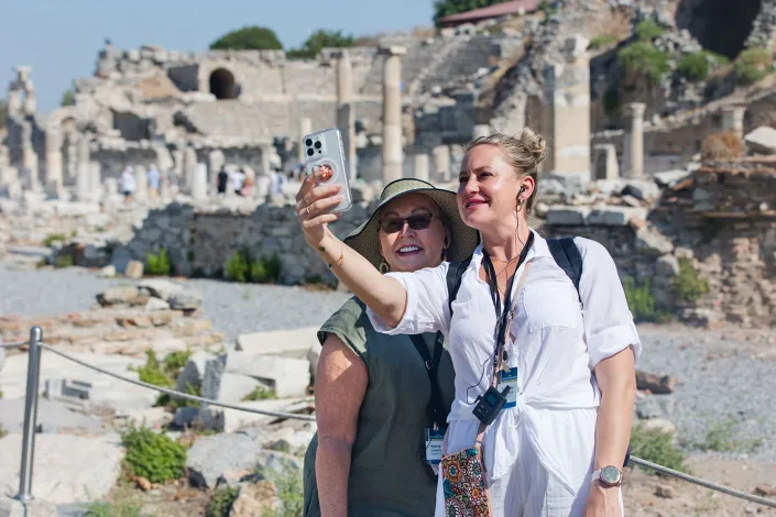 Two travelers smile for a selfie while exploring the ancient ruins of Ephesus, with weathered stone columns and historic structures in the background.