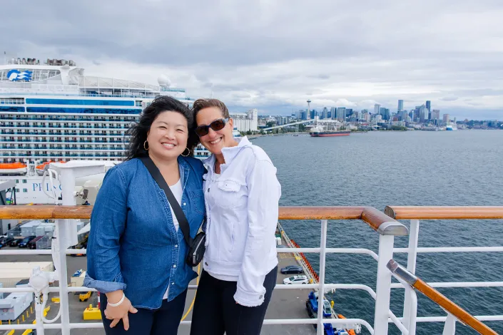 Photo of two women smiling on ship deck, cruise liner and city skyline behind.