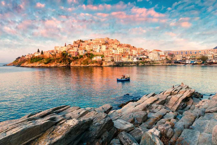Coastal town on hill at sunset, small boat on calm sea, rocks in foreground