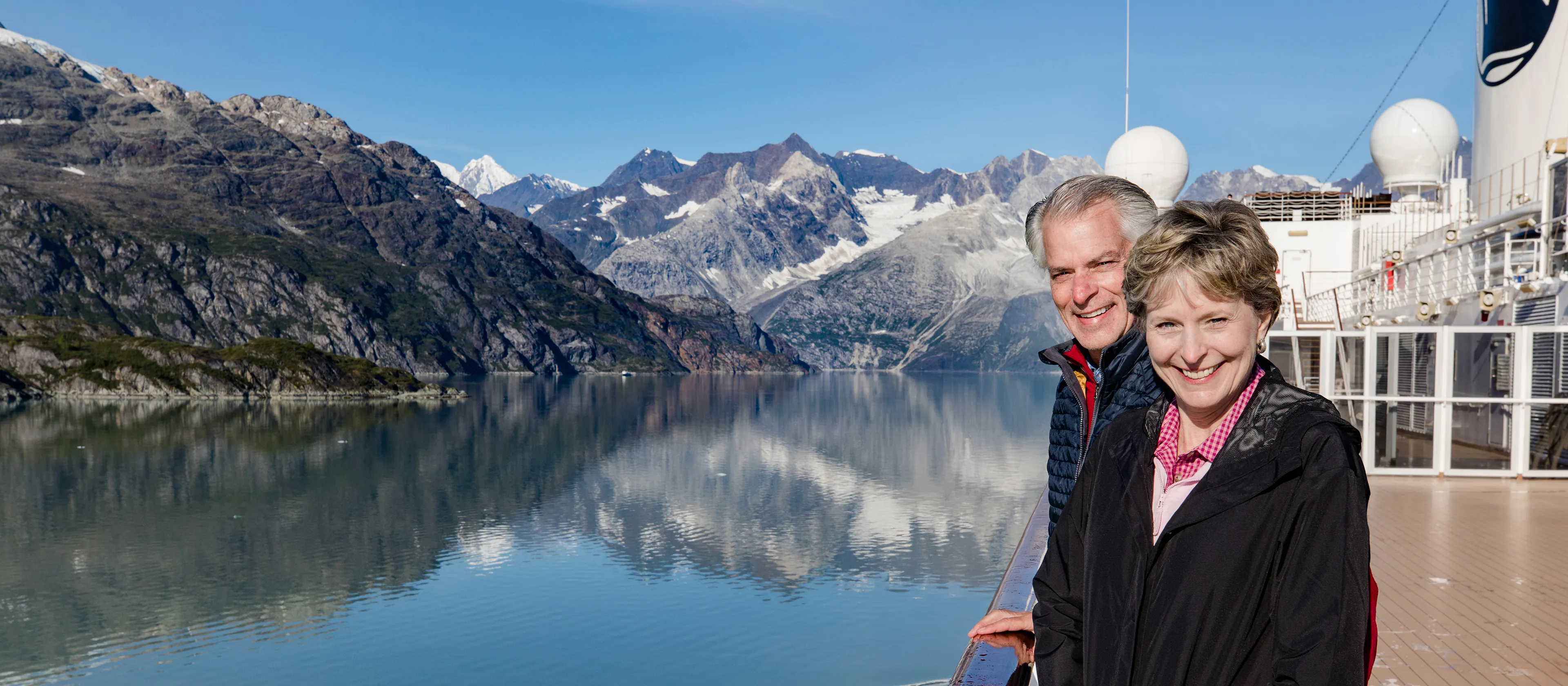 Smiling couple standing on the cruise ship deck with blue water and snowcapped mountains in the background