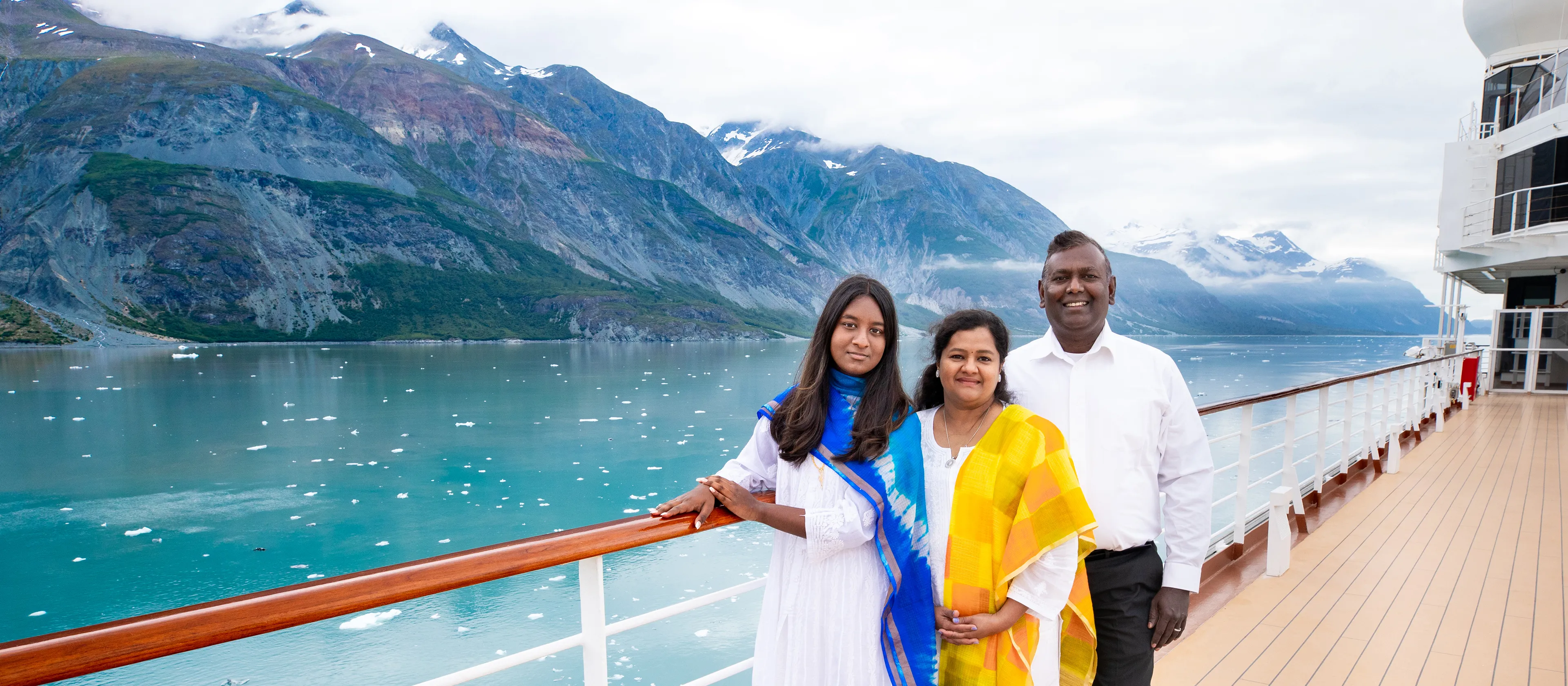 Family of three standing on the cruise ship deck with blue water and snowcapped mountains in the background