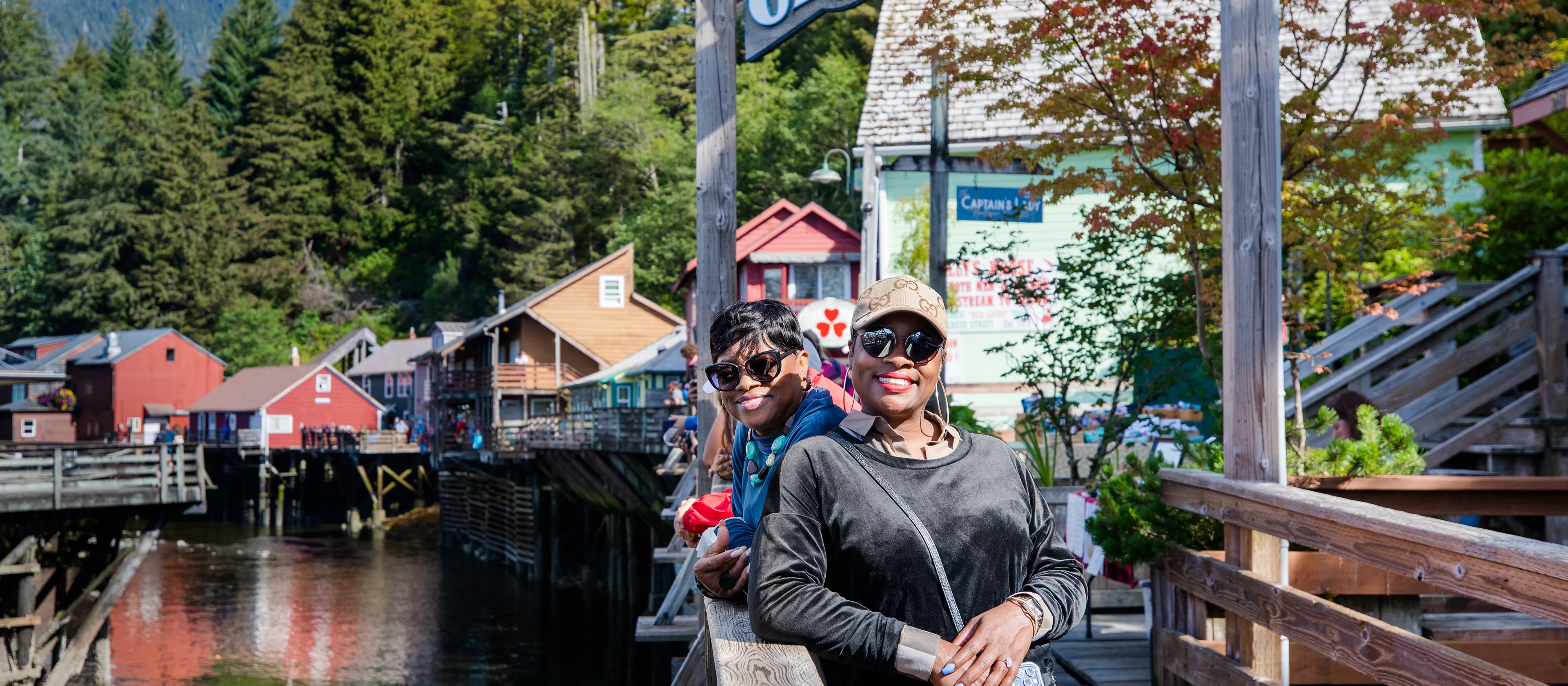 Two smiling women leaning on a wooden railing with trees and colorful buildings in the background