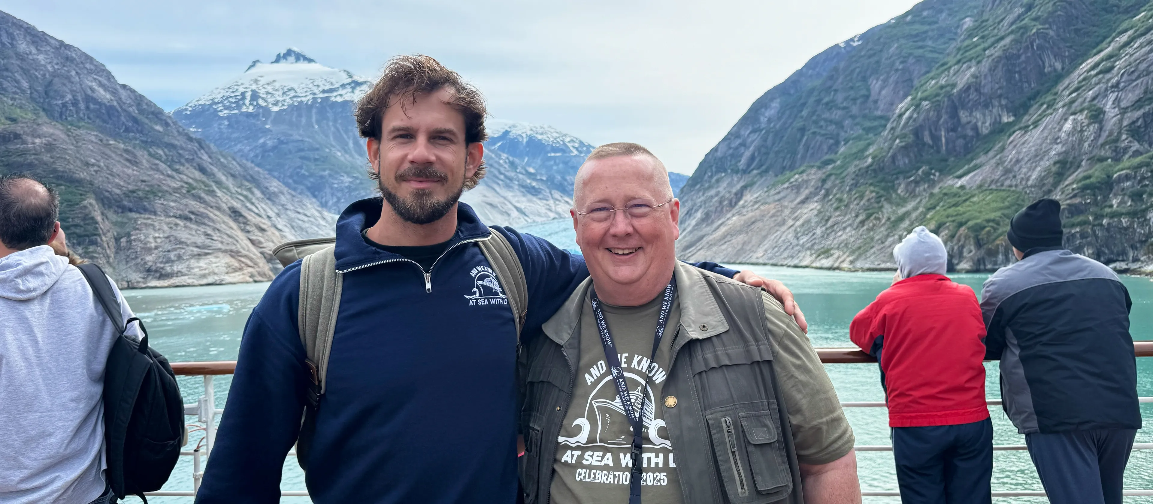 Two men pose together on a cruise ship deck, with a glacier and steep, snow-capped mountains behind them.