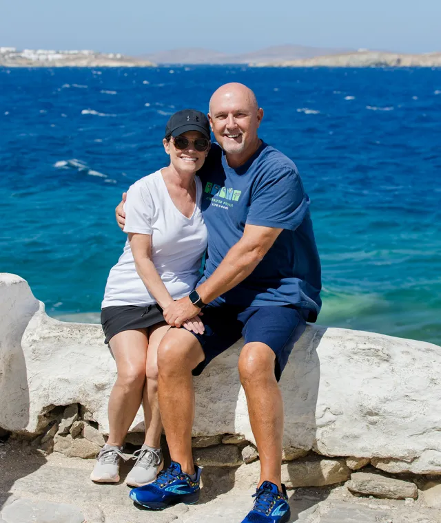 A smiling couple sitting on a stone ledge with the blue ocean behind them