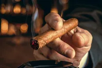 A hand holding a lit cigar over an ashtray in a warmly lit lounge setting