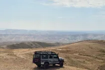 Four-wheel drive vehicle traveling through the Judean Desert near the Dead Sea, surrounded by rolling desert hills beneath a wide, cloud-filled sky.