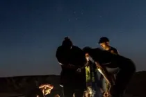 A small group gathered around a light source in the desert under a starry night sky