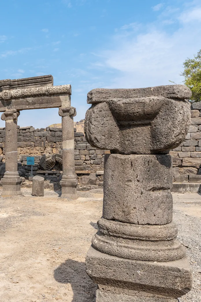 Ancient basalt stone ruins at Chorazin in Galilee, featuring carved columns and archaeological remains of a first-century synagogue beneath a blue sky.