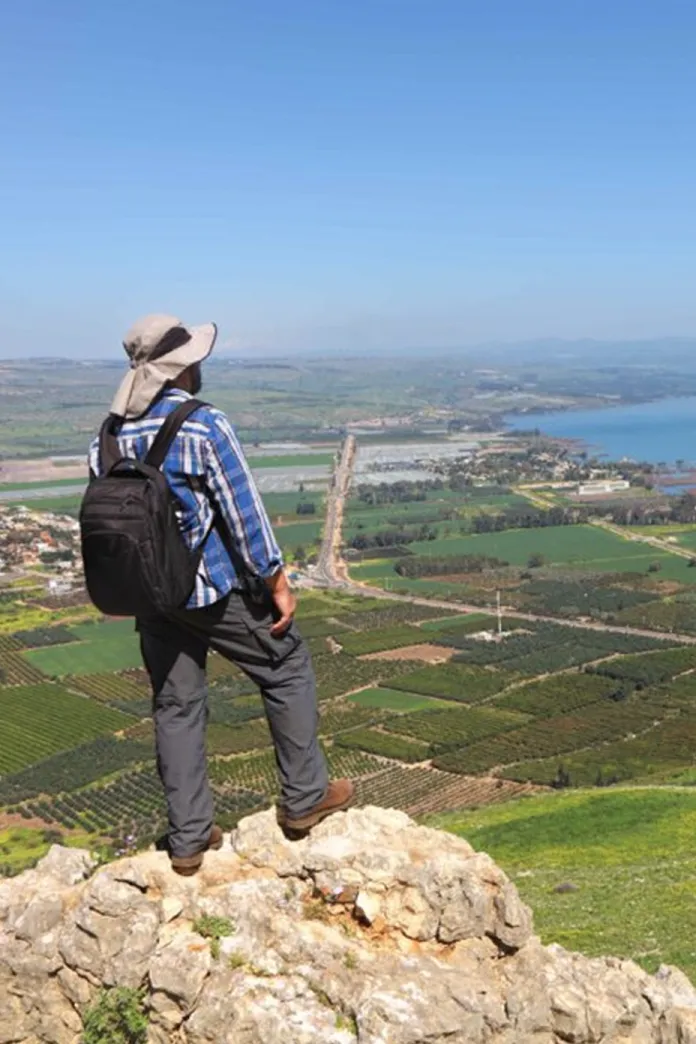 Traveler standing atop Mount Arbel overlooking the Sea of Galilee, with sweeping views of the surrounding countryside, farmland, and shoreline below.